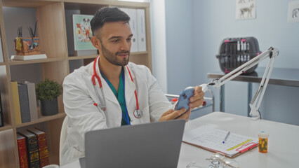 Handsome young hispanic man with a beard in a veterinary clinic, wearing a white lab coat and stethoscope, using a mobile phone while sitting at a desk with a laptop and medical supplies.