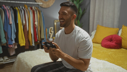 A young hispanic man with a beard smiles while playing video games on a bed in his stylish bedroom with a closet full of colorful clothes.