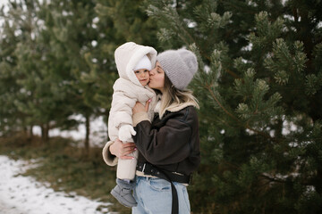 Woman in a brown short sheepskin coat is holding and kissing a baby dressed  in a white overall and hat, surrounded by snow-covered ground and evergreen trees. Joyful moment between mom and baby. 