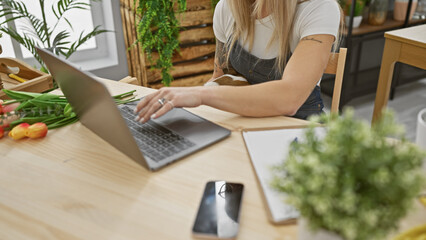 A young woman entrepreneur manages her florist business indoors, using a laptop amid fresh tulips and plants.
