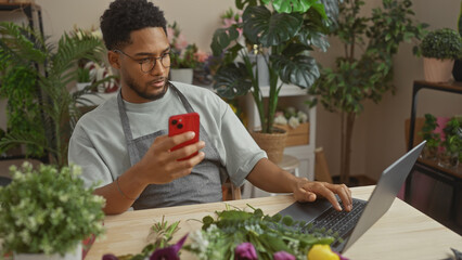 African american man multitasking with smartphone and laptop in a flower shop surrounded by green plants.