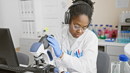 Concentrated african woman scientist working with a microscope in a laboratory setup, wearing headphones