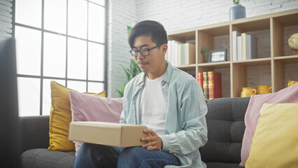 Handsome chinese man relaxing at home unpacking a box in the living room, displaying casual comfort and curiosity.