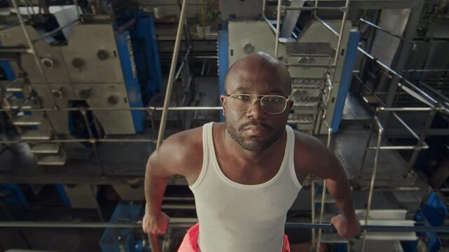 High angle portrait of brutal African American male factory worker in white undershirt leaning back on metal railing and looking at camera with machinery equipment down in background