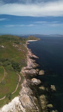 Overlooking the paths and rugged cliffs of Ons Island, the serene sea glistens. Melide Beach and anchored boats appear ahead, with Pontevedra&rsquo;s estuary in the distance under blue sky with high clouds.