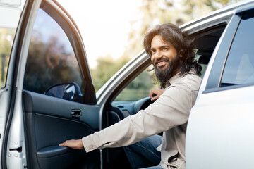 A cheerful man with long hair is seated in the driver's seat of his car, opening the door wide....
