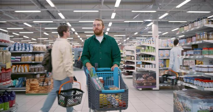 Caucasian Man Strolling with a Cart in a Busy Supermarket Filled with Diverse Shoppers. Families and Solo Customers Picking Groceries. Young Male Filled His Cart with a Variety of Items for the Week