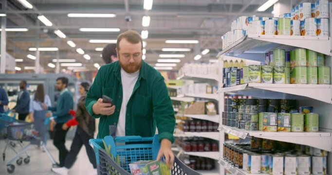 Portrait of a Bearded White Man Using a Smartphone Online Shopping Application while Selecting Canned Goods in a Busy Supermarket, Pushing a Cart Through Well-stocked Aisles