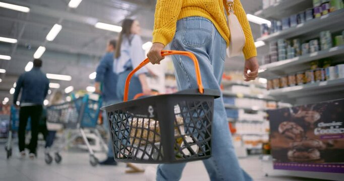 Anonymous Stylish Female Carrying a Shopping Basket Through a Modern Grocery Supermarket, Navigating Aisles Filled with Various Products and Other Shoppers. Low Angle Follow Footage