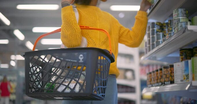 Anonymous Black Female in Yellow Sweater and Jeans Shops for Groceries in a Supermarket, Browsing Shelves and Selecting Items. Shopper Puts Healthy Canned Products in the Basket