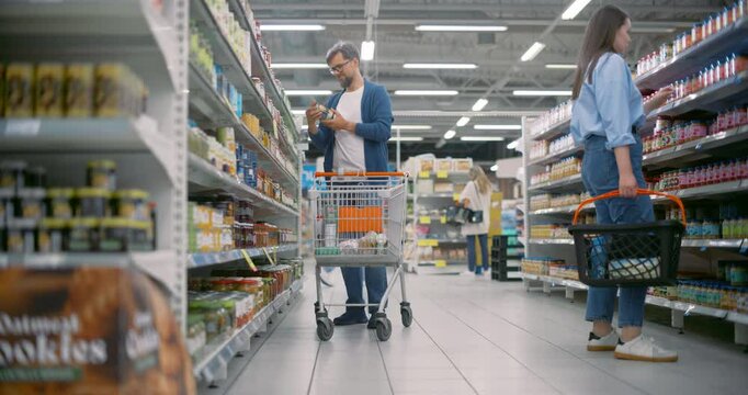 Caucasian Man Wearing Glasses and a Blue Coat Shops in a Modern Supermarket, Examining Canned Beans while Pushing a Cart. Shelves are Stocked With Diverse Products and Shoppers Navigate the Aisles
