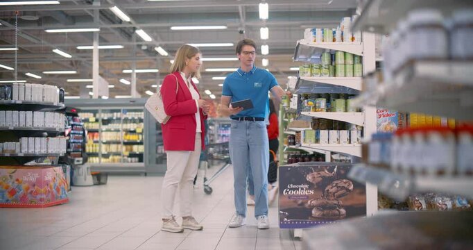 Woman in Red Blazer Receiving Assistance From a Supermarket Manager in a Blue Polo Shirt, Discussing Product Details, Nutritional Value and Discounts. Worker Showing Information on a Tablet Computer