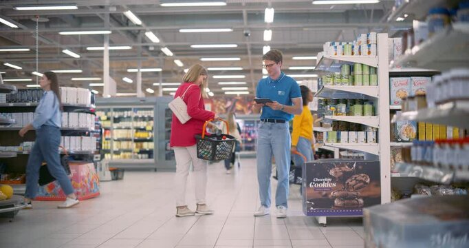 Caucasian Male Supermarket Manager Assists a Young Female Customer, Providing Product Information, Ensuring Excellent Customer Service. Male Organizing Aisle Filled with Various Grocery Items