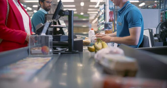 Male Cashier Scanning Product Items for Diverse Customers at a Busy Supermarket Checkout. Bananas, Eggs, Milk are Processed Efficiently, Groceries Rolling Down the Conveyor Belt