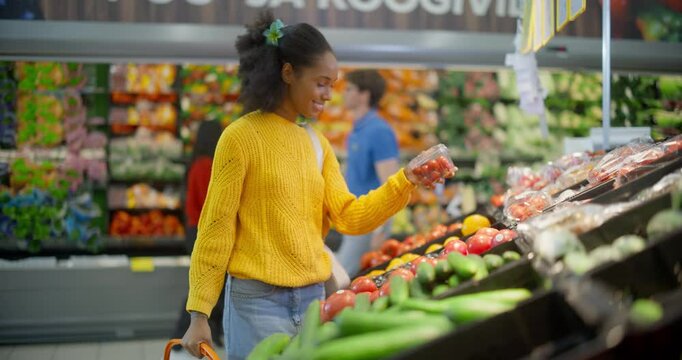 Stylish African American Woman in a Yellow Sweater Shopping for Fresh Produce in a Vibrant Supermarket Aisle, Carefully Selecting Cherry Tomatoes for Their Quality, Freshness and Price