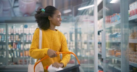 Young Black Woman Standing in Front of the Refrigerated Section, Opening the Fridge to Grab Milk and Eggs. She Quickly Adds Them to Her Basket, Planning Her Next Grocery Choices