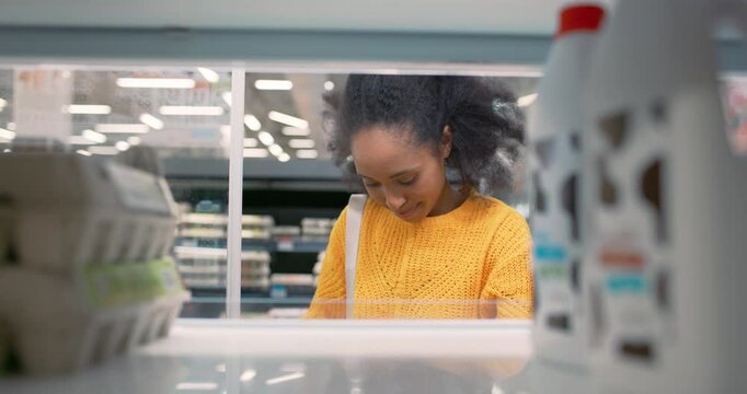 Beautiful Young African American Woman in a Yellow Sweater Takes a Milk Bottle and a Pack of Eggs From a Supermarket Fridge. POV Portrait of a Grocery Shopper From Inside the Fridge