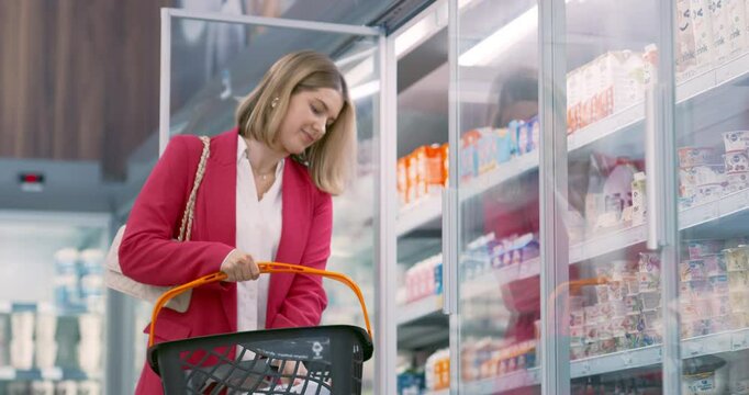 Young Woman Opens the Refrigerator Door in the Grocery Store, Picking Up a Bottle of Milk and a Pack of Eggs. She Adds Them to Her Basket, Choosing Fresh Products for Her Home
