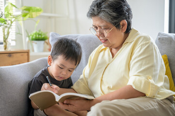 Senior Asian grandmother teaching writing and relaxing with her grandchild at living room happily. Diversity multi generation family activity lifestyle concept. Asian people domestic lifestyle concept