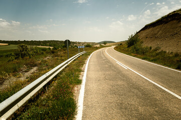 A curving rural road under a clear sky, flanked by greenery and hills, with a visible guardrail and...