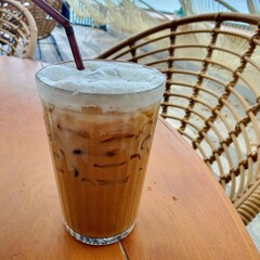 A refreshing iced coffee served in a tall glass, featuring a frothy top and a straw, set against a background of woven chairs.
