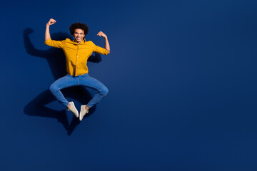 Obraz premium Full body portrait of nice young man jump raise fists flex biceps wear yellow shirt isolated on dark blue color background