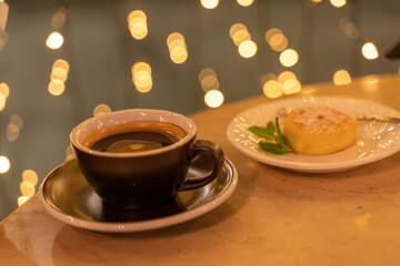 A cup of coffee sits on a table next to a plate of dessert. The dessert is a small cake with a leaf on top. Scene is cozy and inviting, as the coffee and dessert are perfect for a relaxing evening.