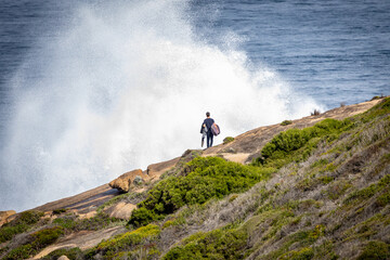 A surfer stands on a rocky coastline watching the huge spray as large waves crash on the rocks in front of him.