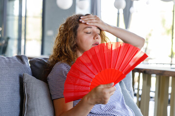 Overheated young woman waving fan suffer from overheating sit on couch at home