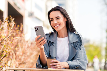 Photo of charming positive woman wear grey trench chatting instagram twitter telegram facebook sitting cafeteria outdoors urban city street