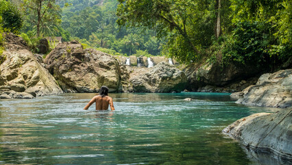 A man swims in a crystal-clear river surrounded by lush tropical forests, rocky cliffs, and...