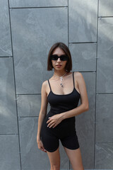 Fashionable beautiful sexy woman with cool sunglasses in trendy fashion black clothes stands on gray background in city