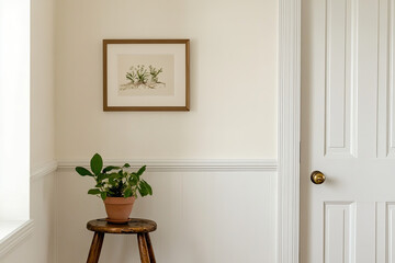 A small framed print on the wall of an apartment, with white walls and a wooden door in front of it, with a potted plant on top of a stool near the door frame, a soft beige background.