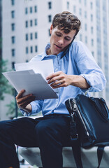 Clever male corporate economist doing accounting with papers during cellphone conversation with operator sitting at urban setting in downtown, formally dressed man communicating with professional
