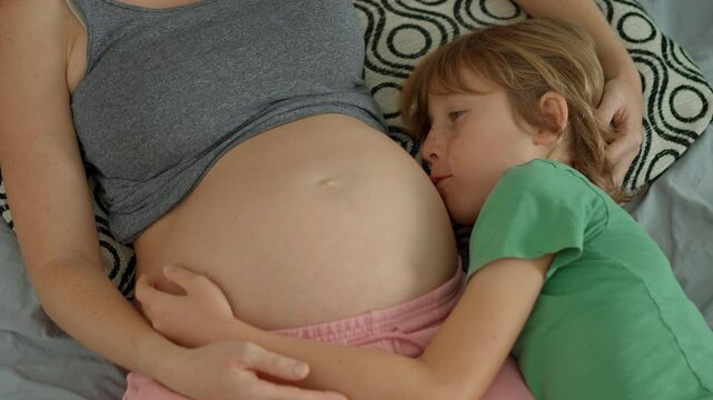 A pregnant woman and her elder son lie on a bed, sharing a tender moment. The boy hugs his mother's belly and talks to his unborn sibling, capturing the beauty of family bonds and the anticipation of