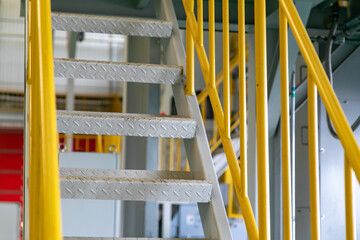 Industrial walkway with yellow handrail and steel structure roof inside the factory, equipped with a staircase secured by handrails.