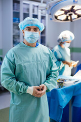 A man in a blue surgical gown stands in front of a surgical table. He is wearing a blue hat and a...