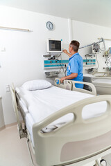 A man in a blue shirt is standing in front of a hospital bed. He is checking the patient's vitals