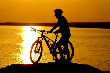 Obraz premium A man is riding a bicycle on a beach at sunset. The water is calm and the sky is orange