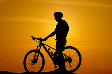 A man is riding a bicycle in the sunset. The man is wearing a helmet and a backpack