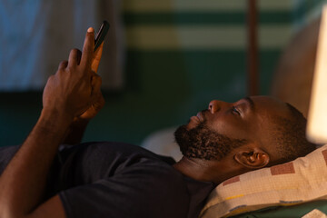 An African man lies in bed, scrolling through his phone in the dim light.