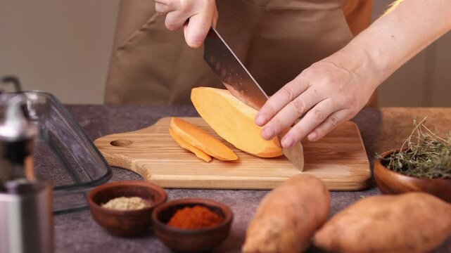 A woman cuts sweet potatoes with a knife to cook them in the oven. Nearby on the table in plates are seasonings, oil, basil. Cooking healthy food at home.