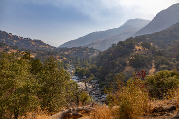 A scenic view of a mountainous valley with a river winding through Sequoia National Park, California. The rugged landscape is dotted with trees, dry hills, and forested slopes under soft sunlight.