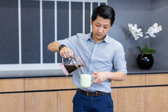 Pouring coffee from French press into mug, Asian man in business attire in office kitchen