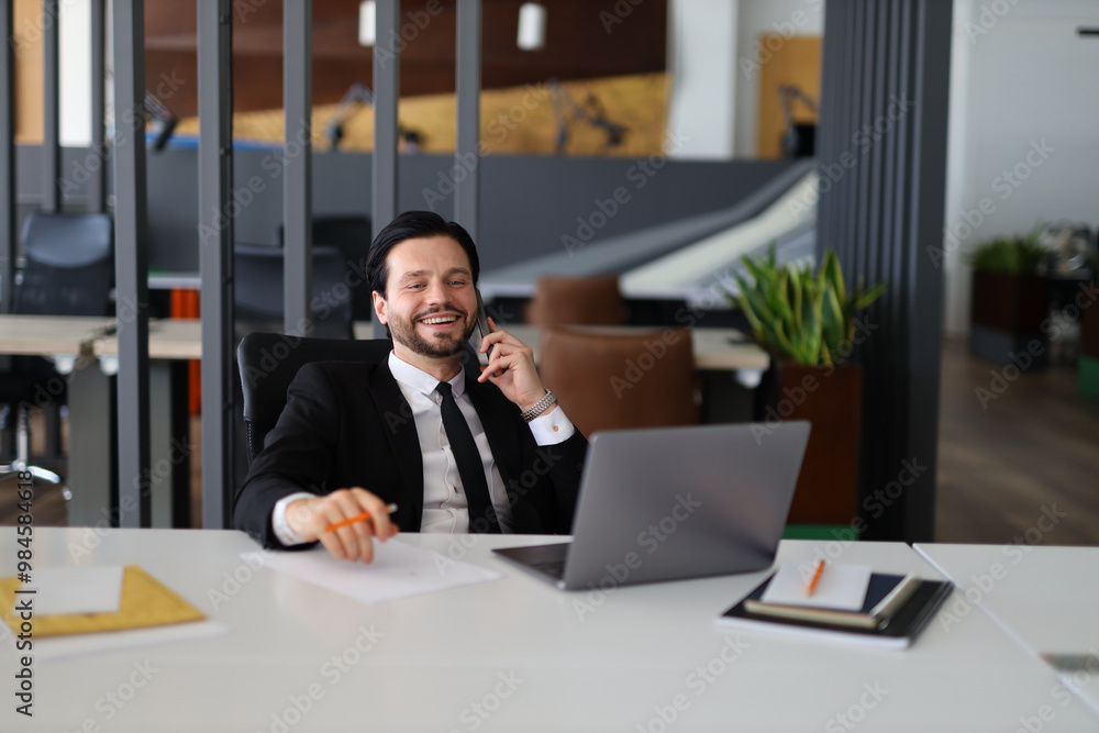 Poster a man in a suit is talking on his cell phone while sitting at a desk. he is smiling and he is in a g