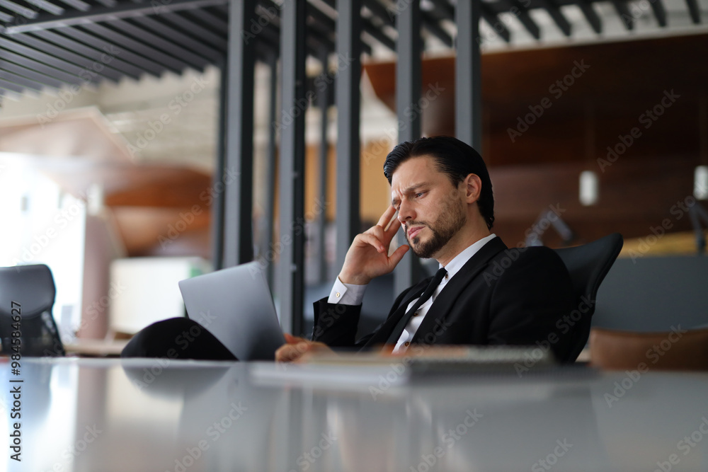 Poster a man in a suit is sitting at a desk with a laptop and a stack of papers. he is looking at the lapto