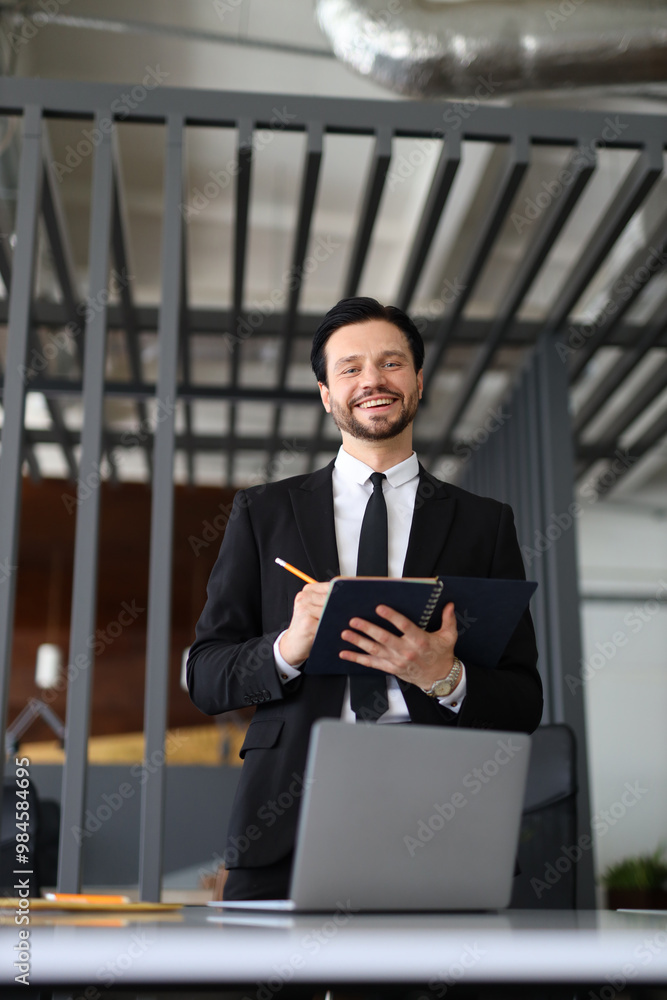 Poster a man in a suit is smiling and holding a notebook and a pencil. he is standing in front of a desk wi