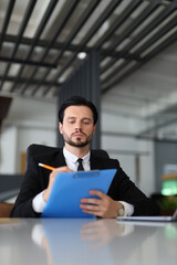 A man in a suit is writing on a blue clipboard. He is focused and serious, likely working on an important project or task