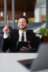 A man in a suit is sitting at a desk with a notebook and a pencil. He is smiling and holding the pencil up in the air. The scene suggests that he is in a creative or problem-solving mindset