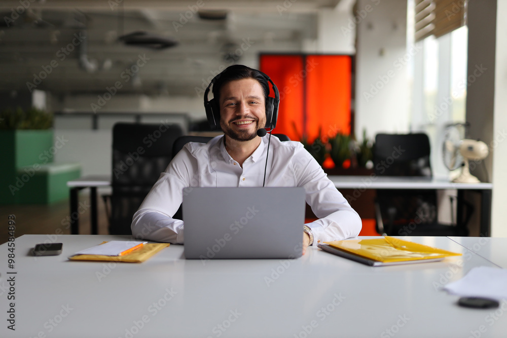Poster a man wearing a headset sits at a desk with a laptop and a stack of papers. he is smiling and he is 